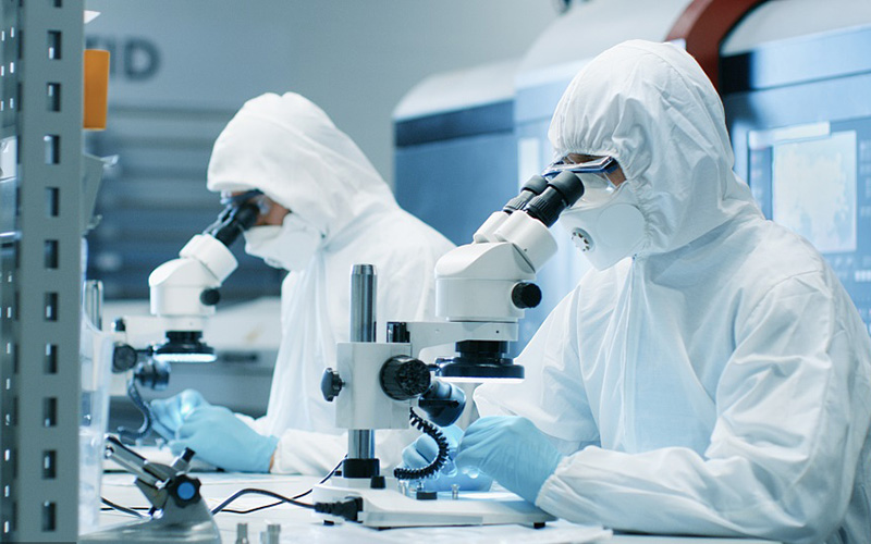 Two Engineers/ Scientists/ Technicians in Sterile Cleanroom Suits Use Microscopes for Component Adjustment and Research. They Work in an Electronic Components Manufacturing Factory.